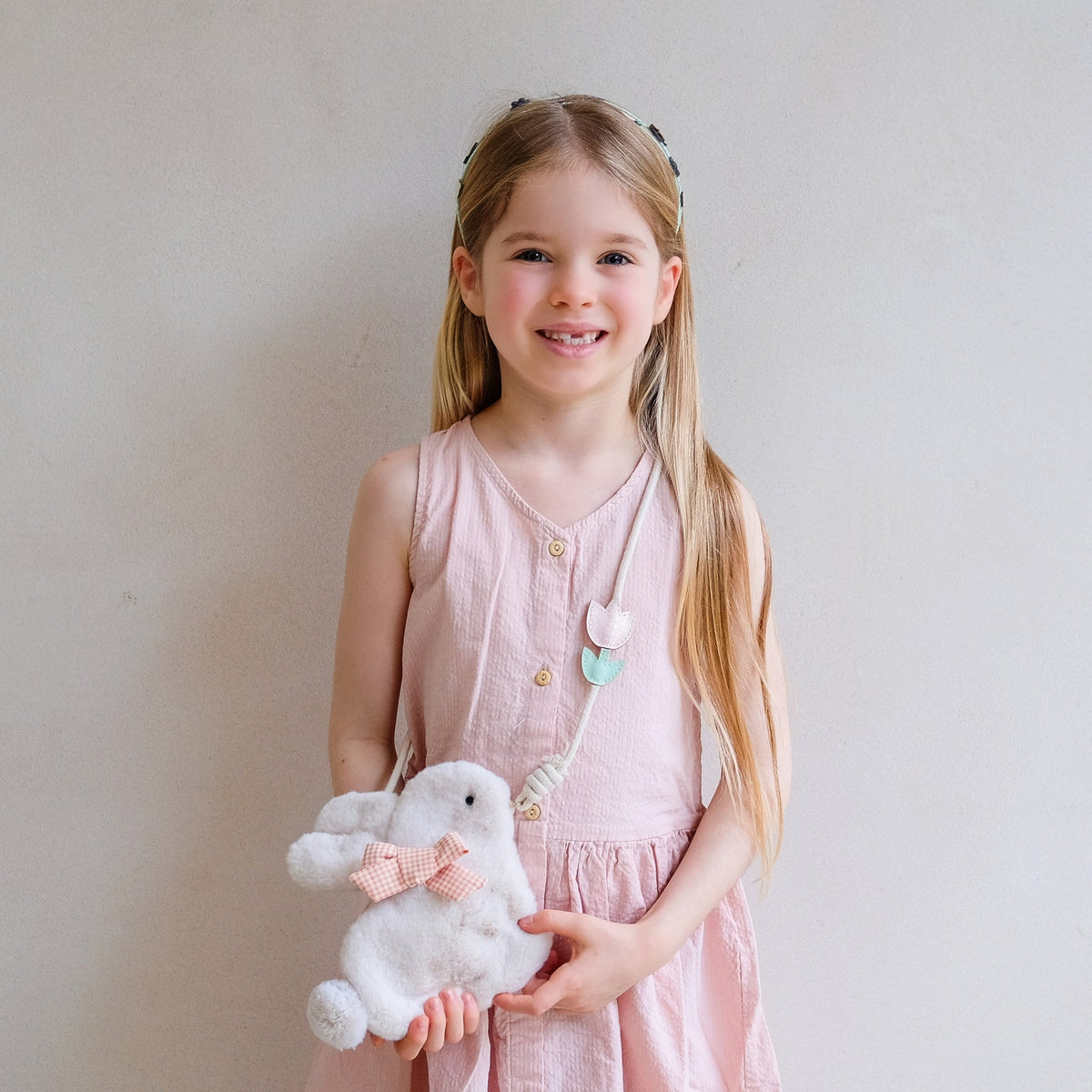 Young girl in a pink dress holding a white teddy bear with a pink bow against a plain background