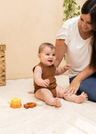 A baby wearing a brown sleeveless romper sits on a cream blanket while a woman in a white t-shirt and blue jeans kneads the baby's legs. Wooden stacking toys are on the blanket, with a wicker basket and a green plant in the background.