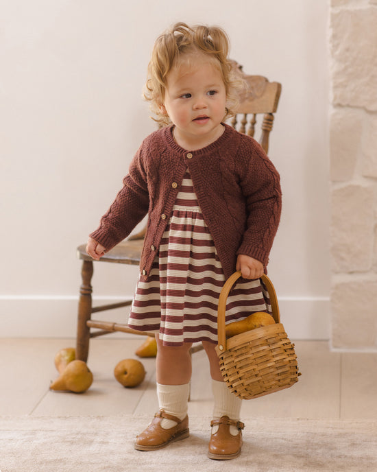 A toddler stands indoors wearing a brown knit cardigan over a red-and-white striped dress, holding a small woven basket. A wooden chair, stone wall, and pears are visible in the background.