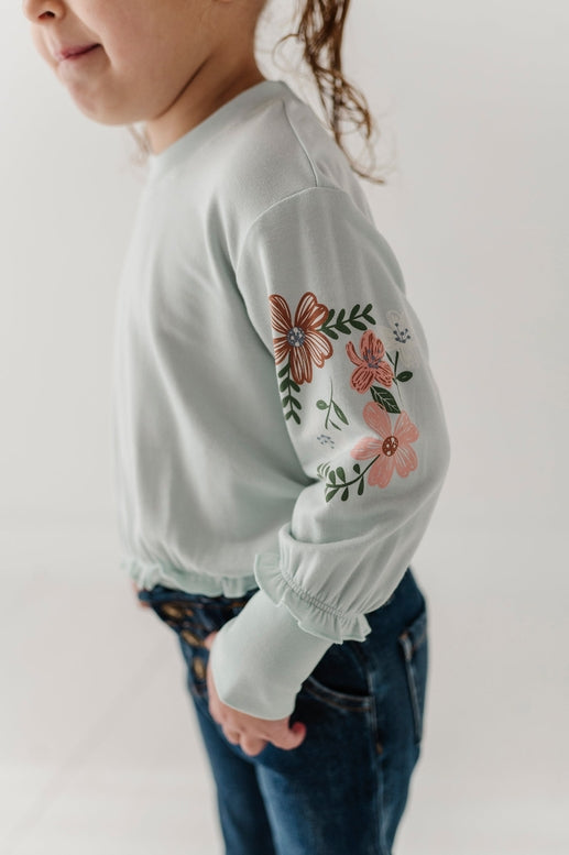 A child wearing a pale mint green long-sleeve top with colorful floral embroidery on the upper sleeve, paired with dark blue jeans, shown against a plain light background.