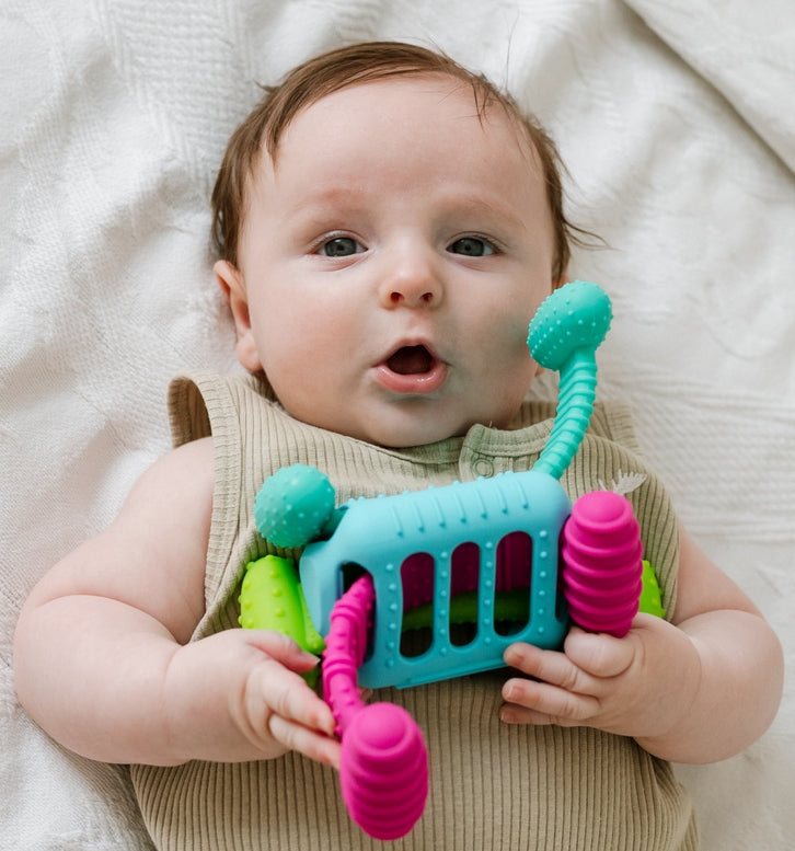 A baby lying on a white textured blanket, holding a colorful sensory toy shaped like a vehicle with a turquoise body, pink wheels, and green bumpers.