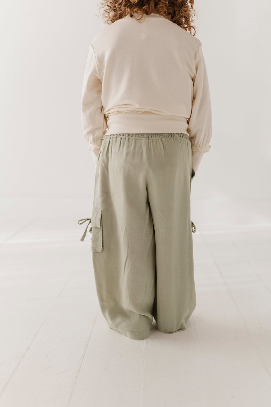 A child viewed from behind wearing a cream sweatshirt and wide-leg sage green cargo pants in a bright, minimalist room.