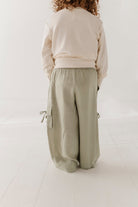 A child viewed from behind wearing a cream sweatshirt and wide-leg sage green cargo pants in a bright, minimalist room.