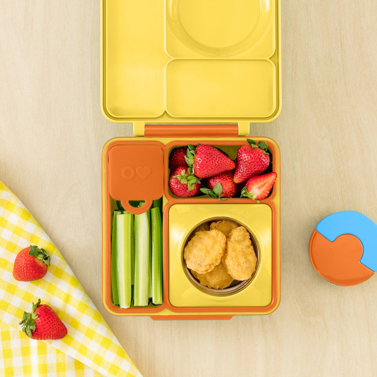 A yellow and orange compartment bento lunch box filled with strawberries, celery sticks, and chicken nuggets, shown on a light wood surface with a matching napkin and lid lid visible.