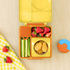 A yellow and orange compartment bento lunch box filled with strawberries, celery sticks, and chicken nuggets, shown on a light wood surface with a matching napkin and lid lid visible.