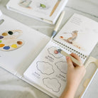 A child's hand holding a paintbrush, actively drawing in a spiral-bound sketchbook. The book features educational watercolor pages, including a paint palette illustration on the left and a squirrel painting on the right.