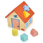 A wooden house shape sorter with its red roof panel detached and laid flat, revealing the interior while five colorful wooden shapes—a mint green cube, yellow cylinder and triangle, pink heart, and light blue cross—sit in front of the toy.