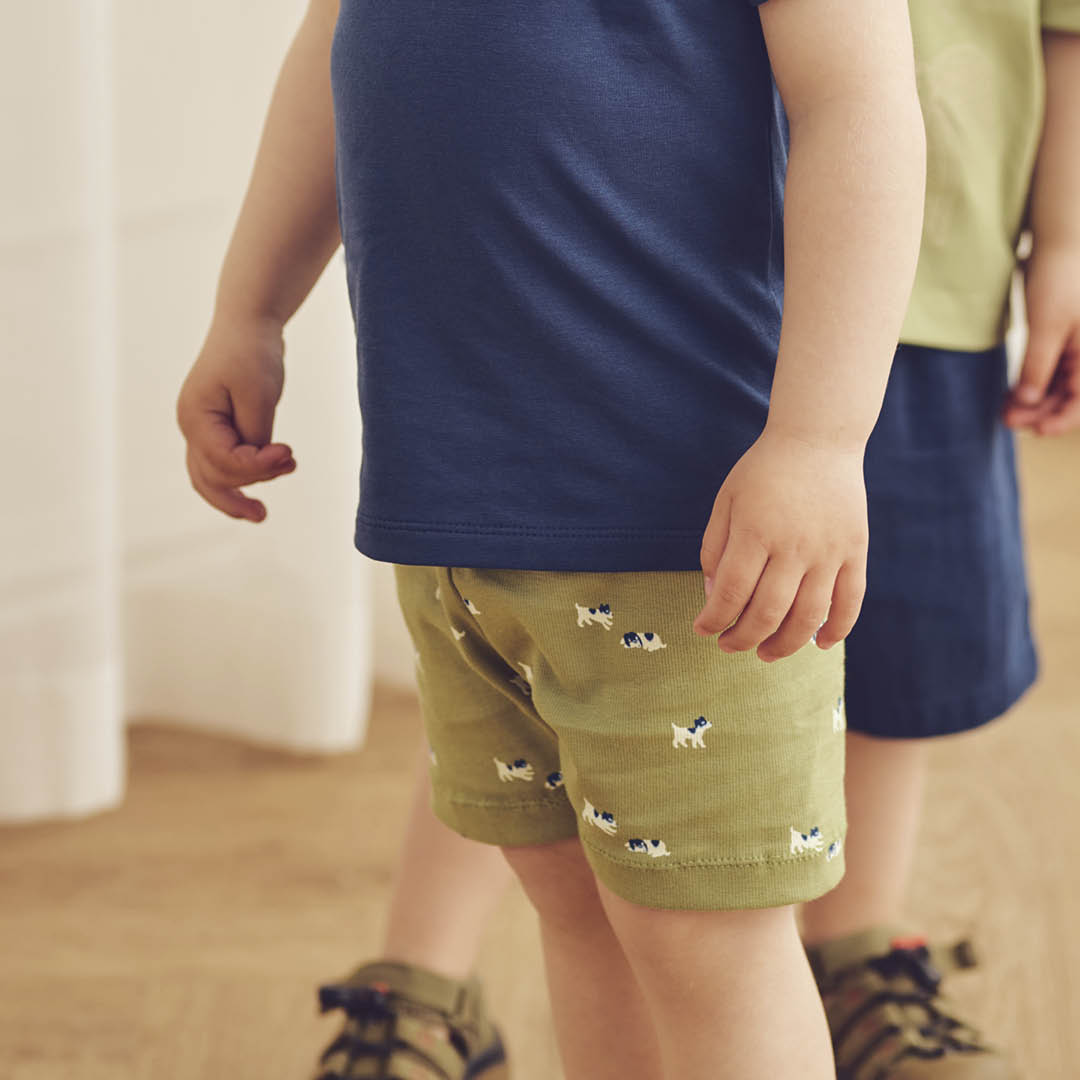 Children wearing navy shirts and green shorts with dog prints on a wooden floor.