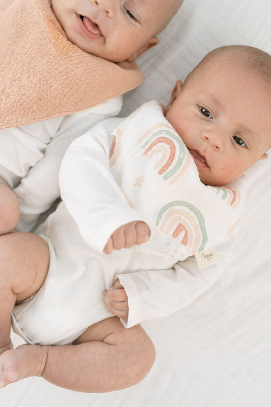 A baby wearing a white long-sleeve bodysuit with a pastel rainbow print on the chest, lying on a white blanket next to another baby with a peach bib.