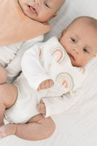 A baby wearing a white long-sleeve bodysuit with a pastel rainbow print on the chest, lying on a white blanket next to another baby with a peach bib.