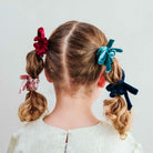 A child viewed from the back wearing three velvet hair bows in red, teal, and navy, attached to pigtails.