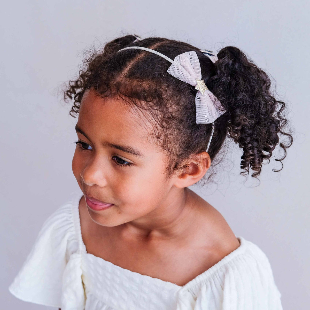 A young girl with curly dark hair wearing a white bow headband, styled in a half-up ponytail, looking down and to the side. She is wearing a white off-the-shoulder top against a plain light gray background.