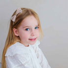 A young girl with long blonde hair wearing a light pink bow headband and a white eyelet blouse, photographed against a plain light gray background.
