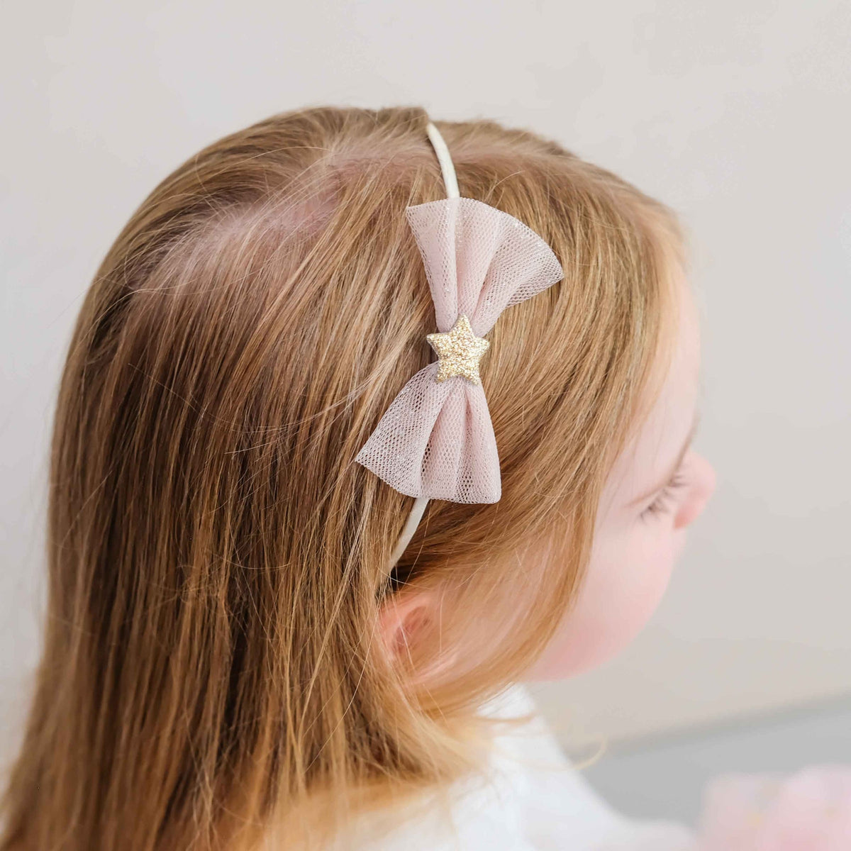 A young girl with light brown hair is shown in profile wearing a pale pink mesh bow headband with a glittery gold star accent.