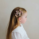 A side-profile view of a young girl with long light brown hair wearing six small bow-shaped hair clips in soft pink, beige, and cream tones against a plain light gray background.