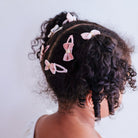 Pink bow hair clips with gold center accents styled in a child's curly dark hair, shown from a side-back angle against a light gray background.