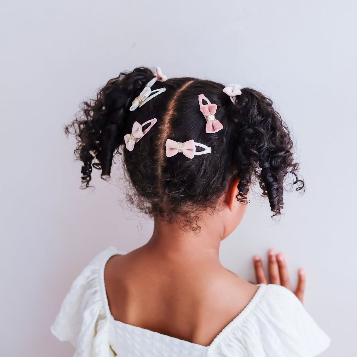 A child seen from behind with curly dark hair styled in two pigtails, wearing several small bow-shaped hair clips in pale pink, white, and light blue against a plain light background.