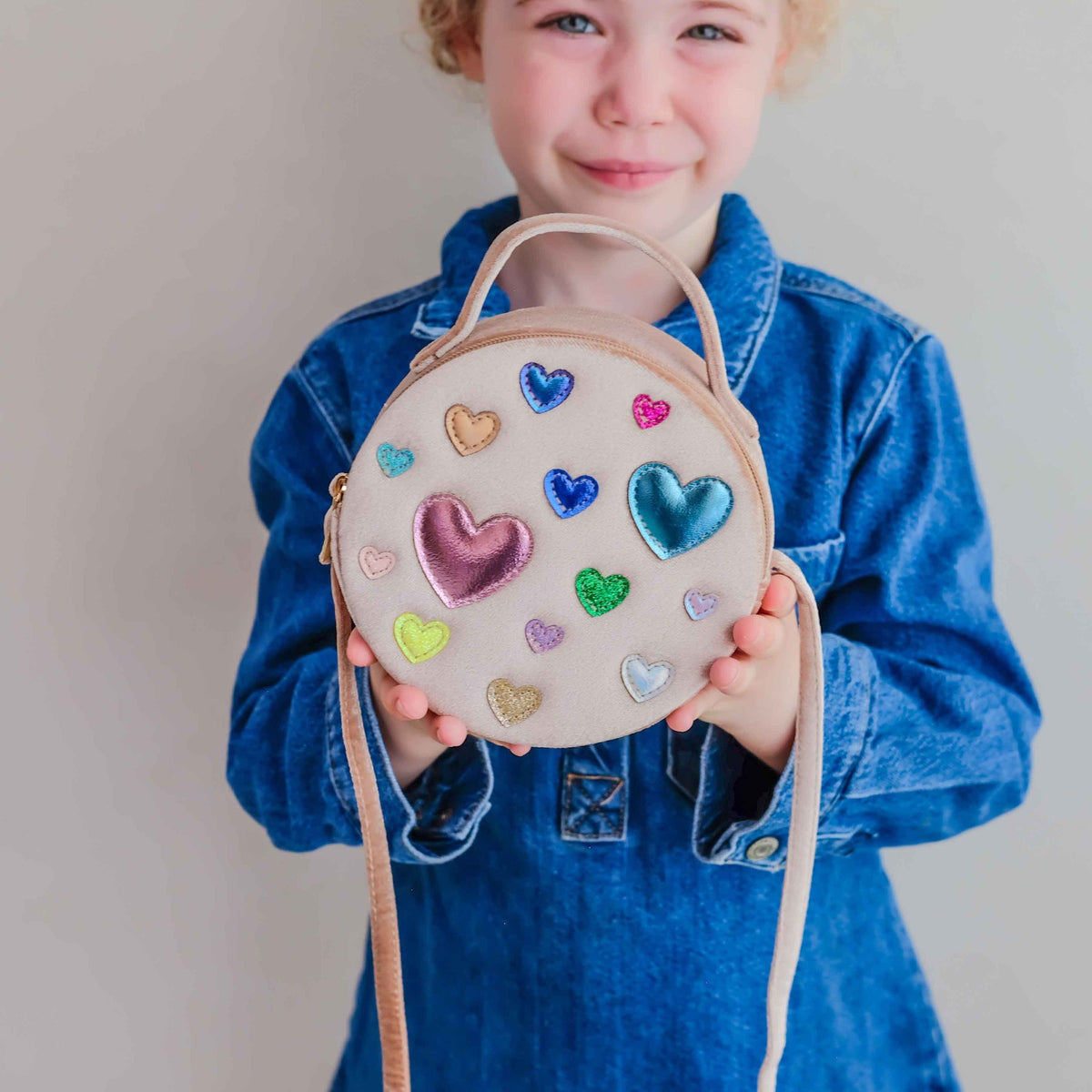 A young child wearing a blue denim jacket holds a round beige fabric crossbody bag decorated with colorful embroidered hearts. The child is smiling and looking at the camera against a plain light gray background.