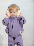 A toddler wearing a matching lavender penguin print zip-up jacket and pants set, holding white earmuffs with a blue headband. The jacket features black penguin graphics and a gold zipper.