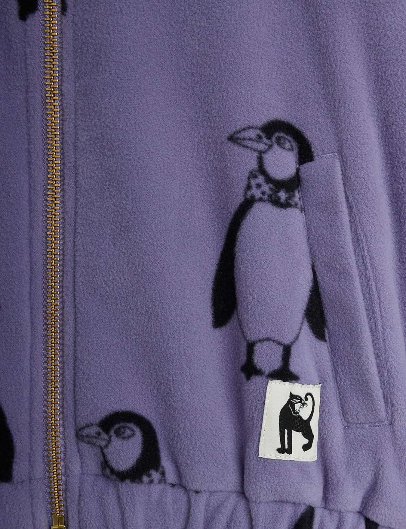 Close-up of a lavender fleece zip-up jacket featuring a black penguin print and a small white brand patch with a black cat silhouette.