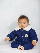 A baby wearing a royal blue quilted baby snowsuit with a white faux-fur collar, gold snap buttons, striped cuffs and hem, and a cartoon patch on the chest.