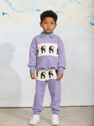 A young boy modeling a lavender two-piece tracksuit with a white chest and waist panel featuring black dog illustrations, a light blue collar, and light blue cuffs, paired with white sneakers.