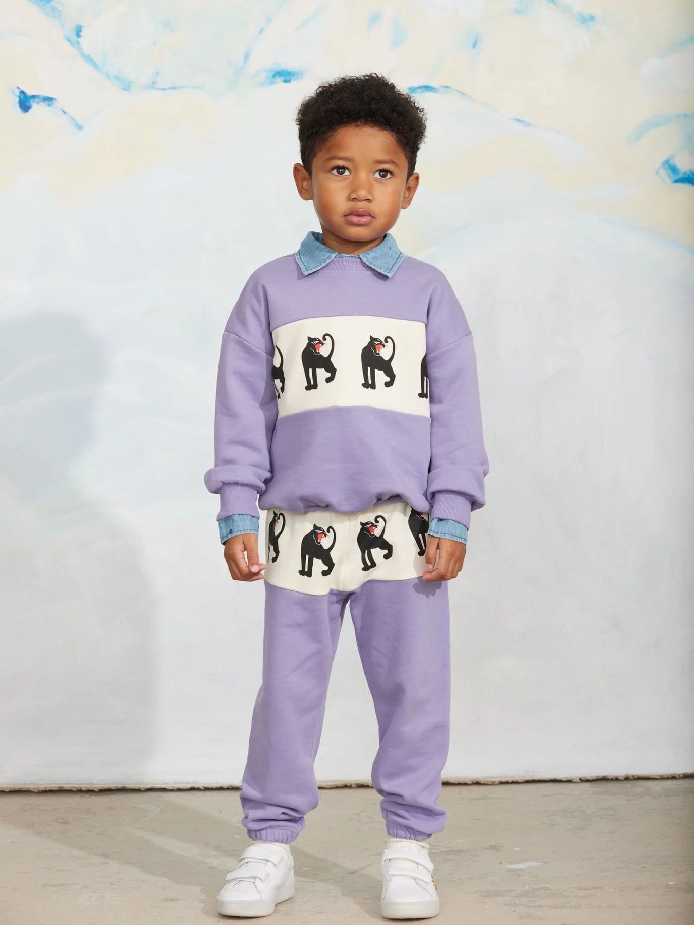 A young child modeling a lavender two-piece tracksuit with a white chest panel featuring black dog illustrations and light blue collar/cuff accents, paired with white sneakers, against a light textured wall.