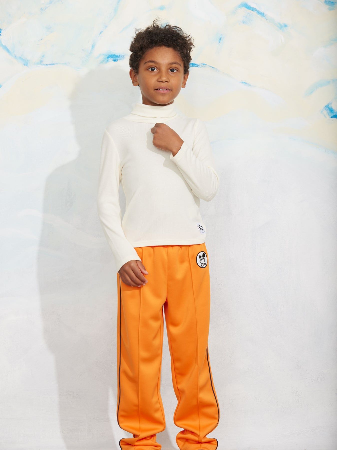 A child model wearing a white long-sleeve mock neck top and bright orange track pants with black side stripes and a circular logo patch, standing against a white textured backdrop.