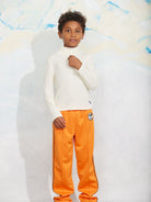 A child model wearing a white long-sleeve mock neck top and bright orange track pants with black side stripes and a circular logo patch, standing against a white textured backdrop.