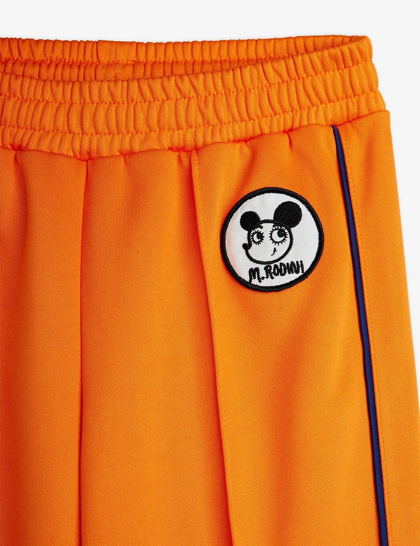 Close-up of bright orange athletic shorts with an elastic waistband, a circular black-and-white cartoon patch reading 'M.ROOMY' on the left side, and a thin dark blue side stripe.