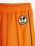 Close-up of bright orange athletic shorts with an elastic waistband, a circular black-and-white cartoon patch reading 'M.ROOMY' on the left side, and a thin dark blue side stripe.
