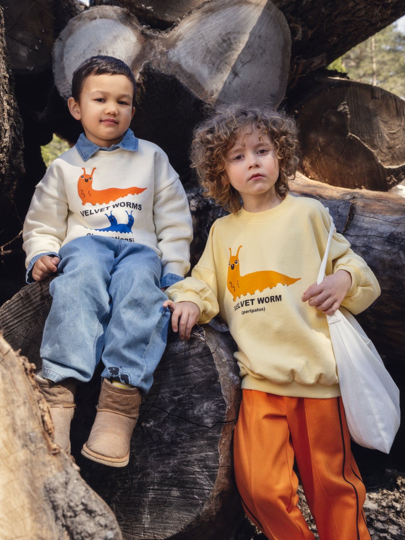 Two children sitting and standing outdoors among large tree logs, wearing matching cream and pale yellow sweatshirts with an orange worm graphic and the text 'VELVET WORM'.