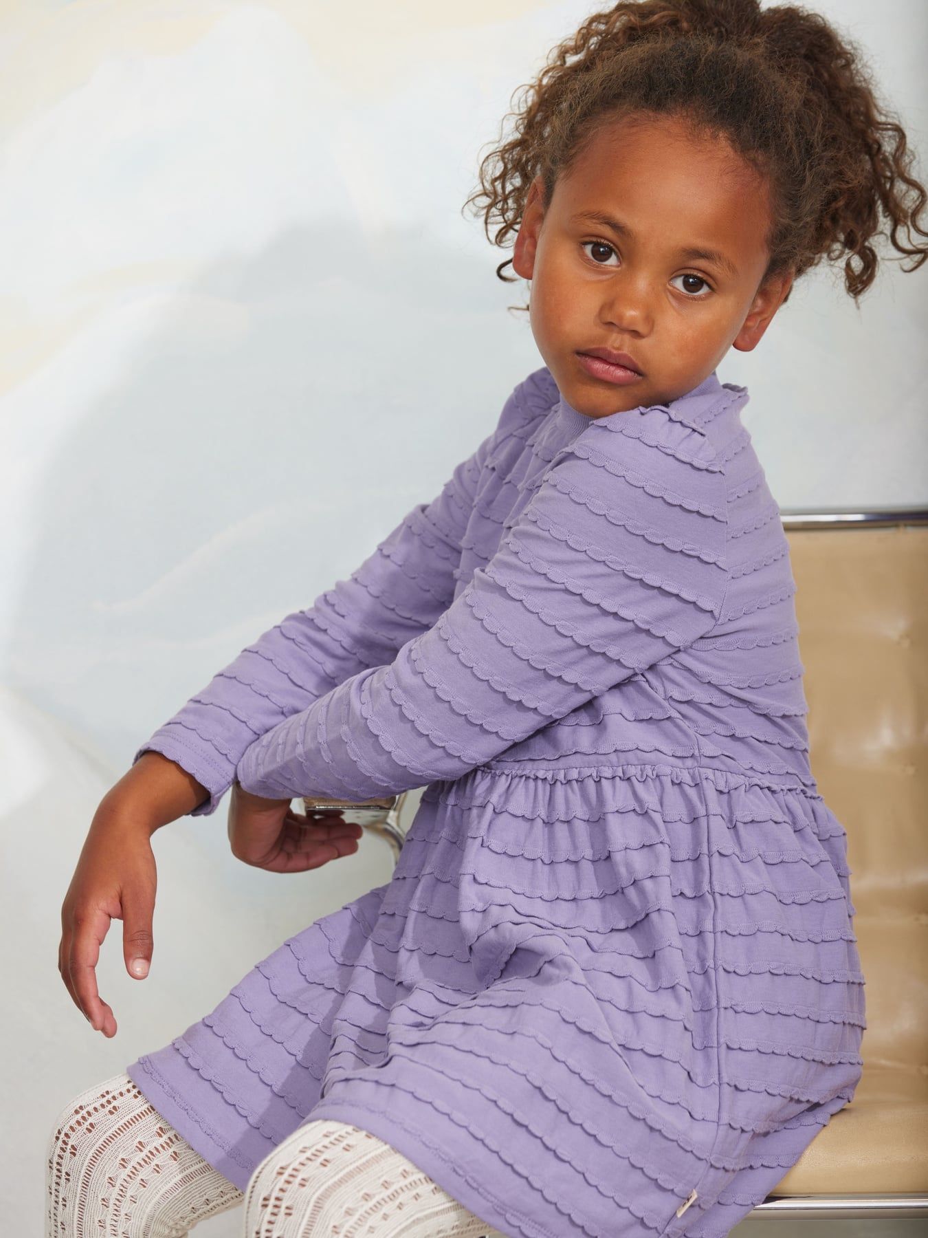 A young girl with curly brown hair wearing a lavender purple textured long-sleeve dress and white knit tights, sitting on a beige chair against a light background.