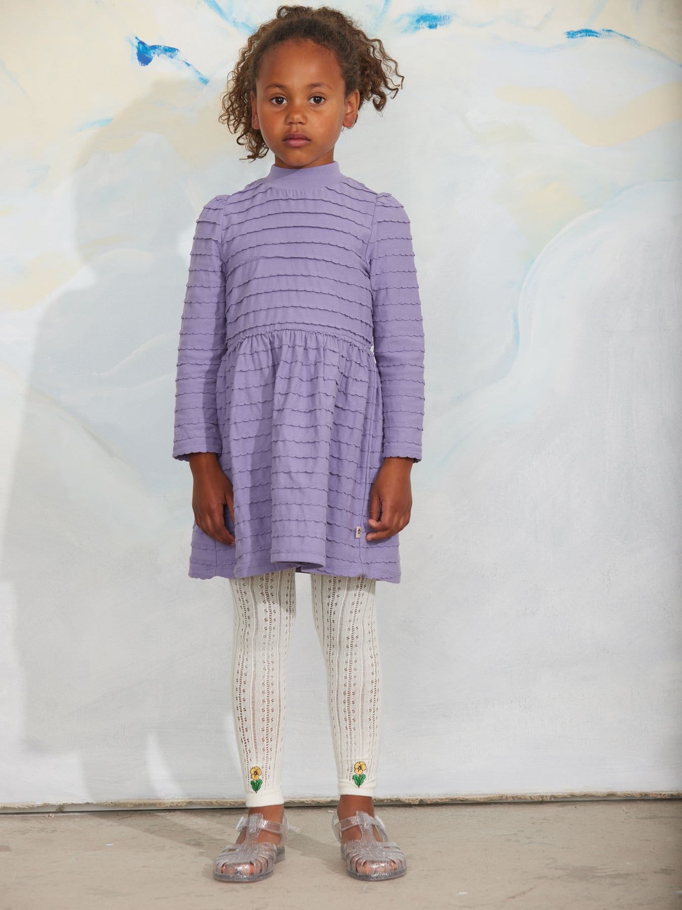 A young girl with curly brown hair wearing a lavender purple textured long-sleeve dress and white knit tights in a white background.