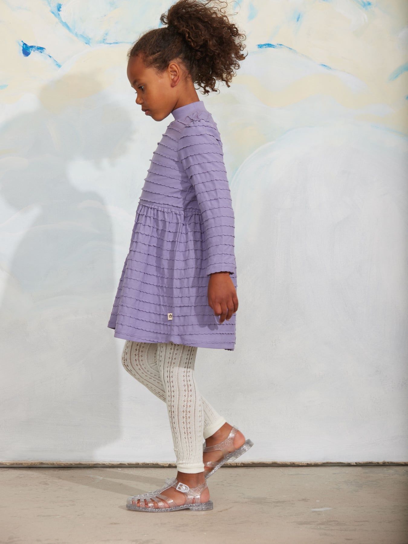 A young girl with curly brown hair wearing a lavender purple textured long-sleeve dress and white knit tights.