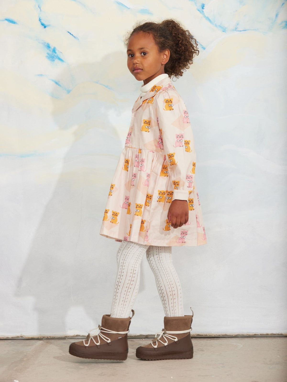 A young girl stands against a painted backdrop wearing a long-sleeve cream dress with a yellow and pink animal print, white knit tights, and brown lace-up boots.
