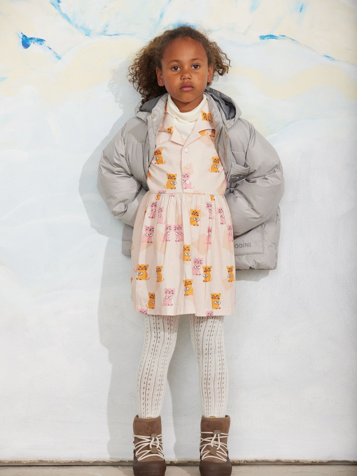 A young girl stands against a white wall wearing a light pink dress with a yellow and orange bear print, a gray puffer jacket, white knit tights, and brown lace-up boots.