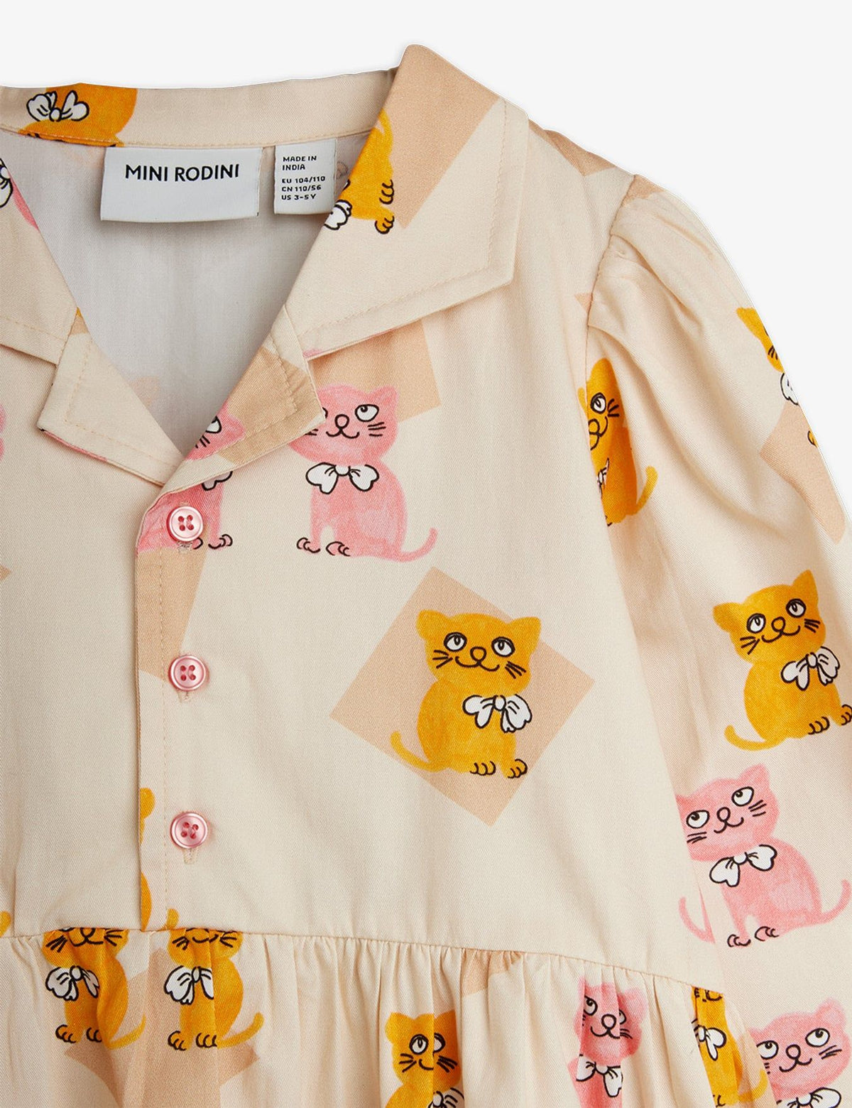 Close-up of a baby dress by Mini Rodini, featuring a cream-colored fabric with a playful print of yellow and pink cartoon cats, a buttoned collar, and pink buttons.