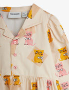 Close-up of a baby dress by Mini Rodini, featuring a cream-colored fabric with a playful print of yellow and pink cartoon cats, a buttoned collar, and pink buttons.
