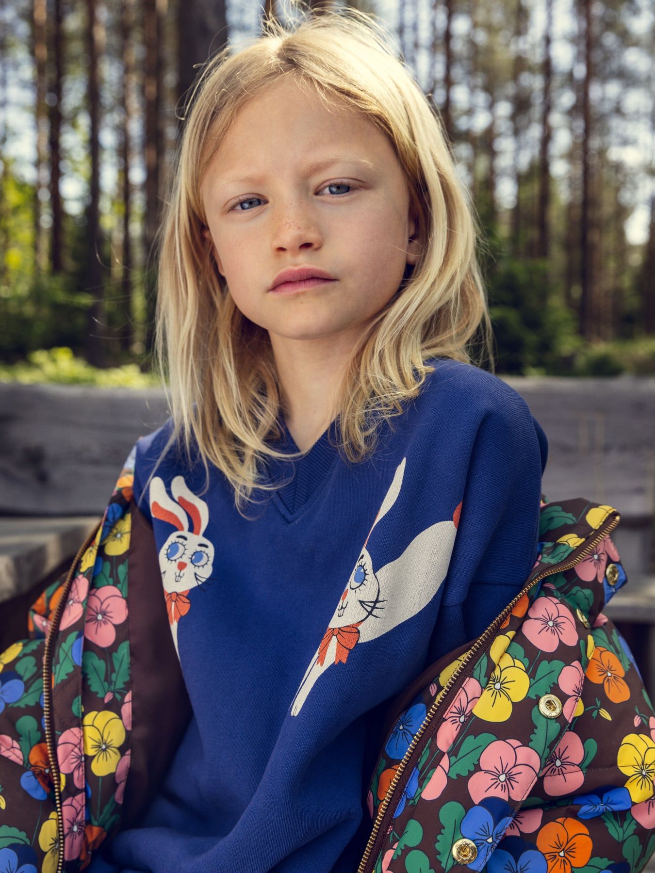 A young blonde girl wearing a blue sweatshirt with a rabbit graphic and a colorful floral zip-up jacket, sitting outdoors in front of a blurred forest background.