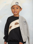 A young boy wearing a black and beige sweatshirt with mouse graphics, a light gray knit cardigan, and a cream beanie with a panda logo. The cardigan features a matching circular panda patch on the sleeve.