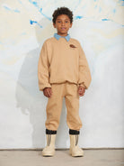 A child model wearing a beige sweatshirt and matching sweatpants with a light blue collar detail, black ankle cuffs, and beige boots, standing against a textured white wall.