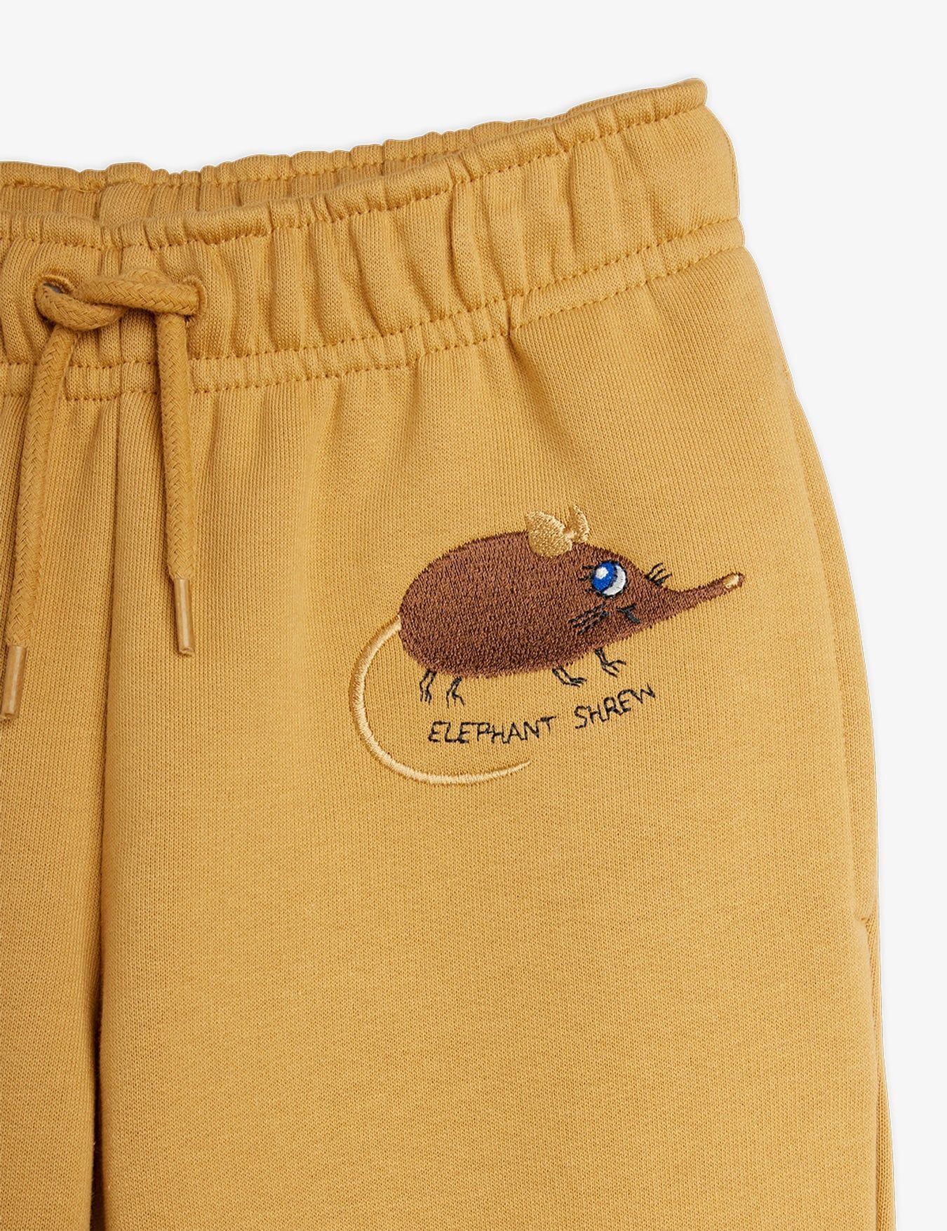 Close-up of mustard yellow sweatpants featuring a brown embroidered elephant shrew on the left leg with the text 'ELEPHANT SHREN' beneath it, shown against a plain white background.