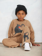 A child wearing a beige knit two-piece outfit with black panther graphics, a black ribbed beanie, and white sneakers, sitting against a light gray wall.