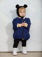 A toddler stands against a white wall wearing a navy blue quilted puffer jacket with a black hood featuring mouse ears, cream ribbed cuffs, black pants, and white sneakers.