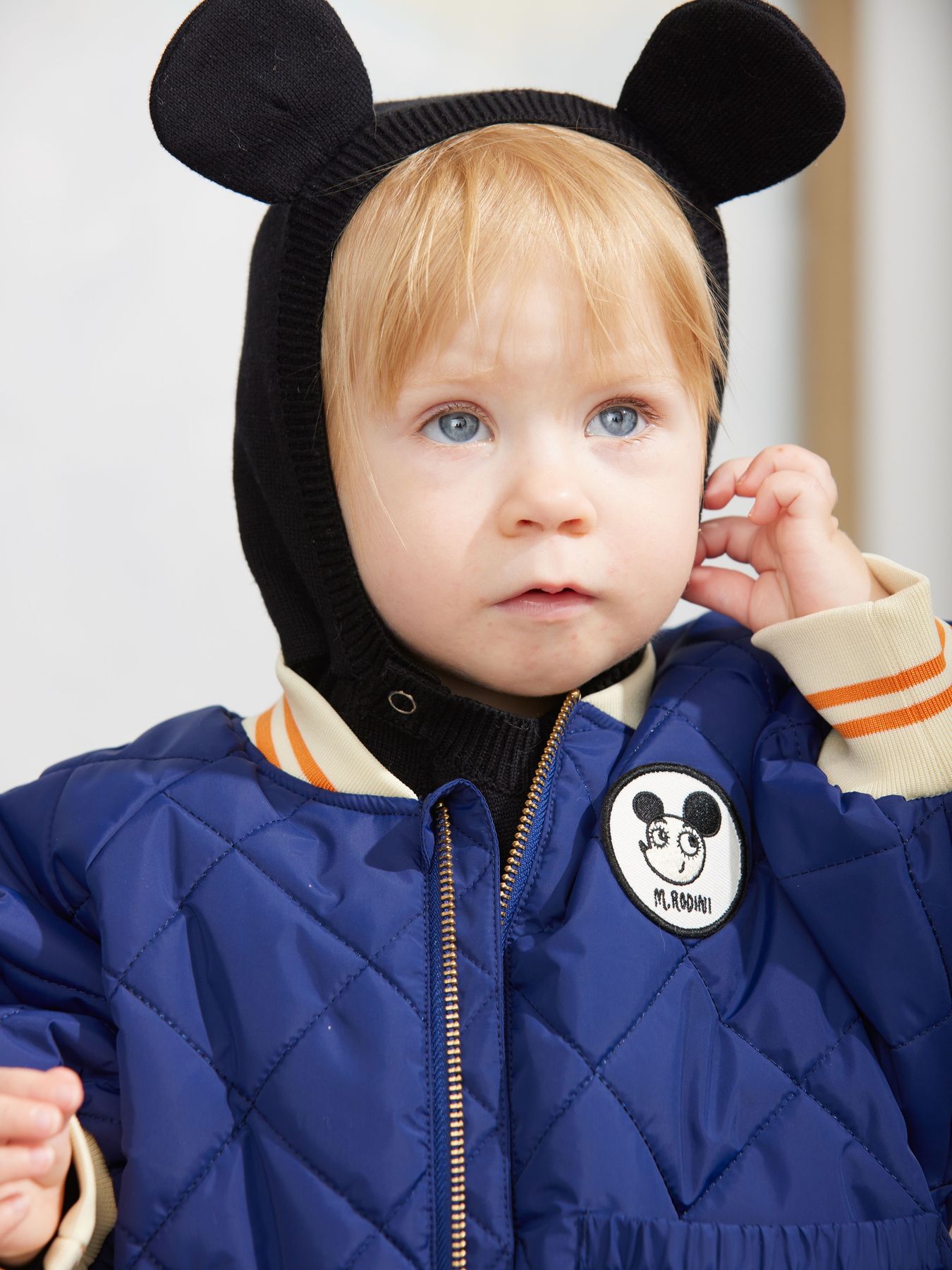 A toddler wearing a navy blue quilted puffer jacket with a circular Mickey Mouse patch on the chest, a black knit hood with mouse ears, and striped cream sleeves, photographed against a soft light background.