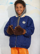A smiling young boy wearing a blue quilted puffer jacket with a gray hood, brown fuzzy clawed gloves, and bright orange pants, standing against a light textured wall.