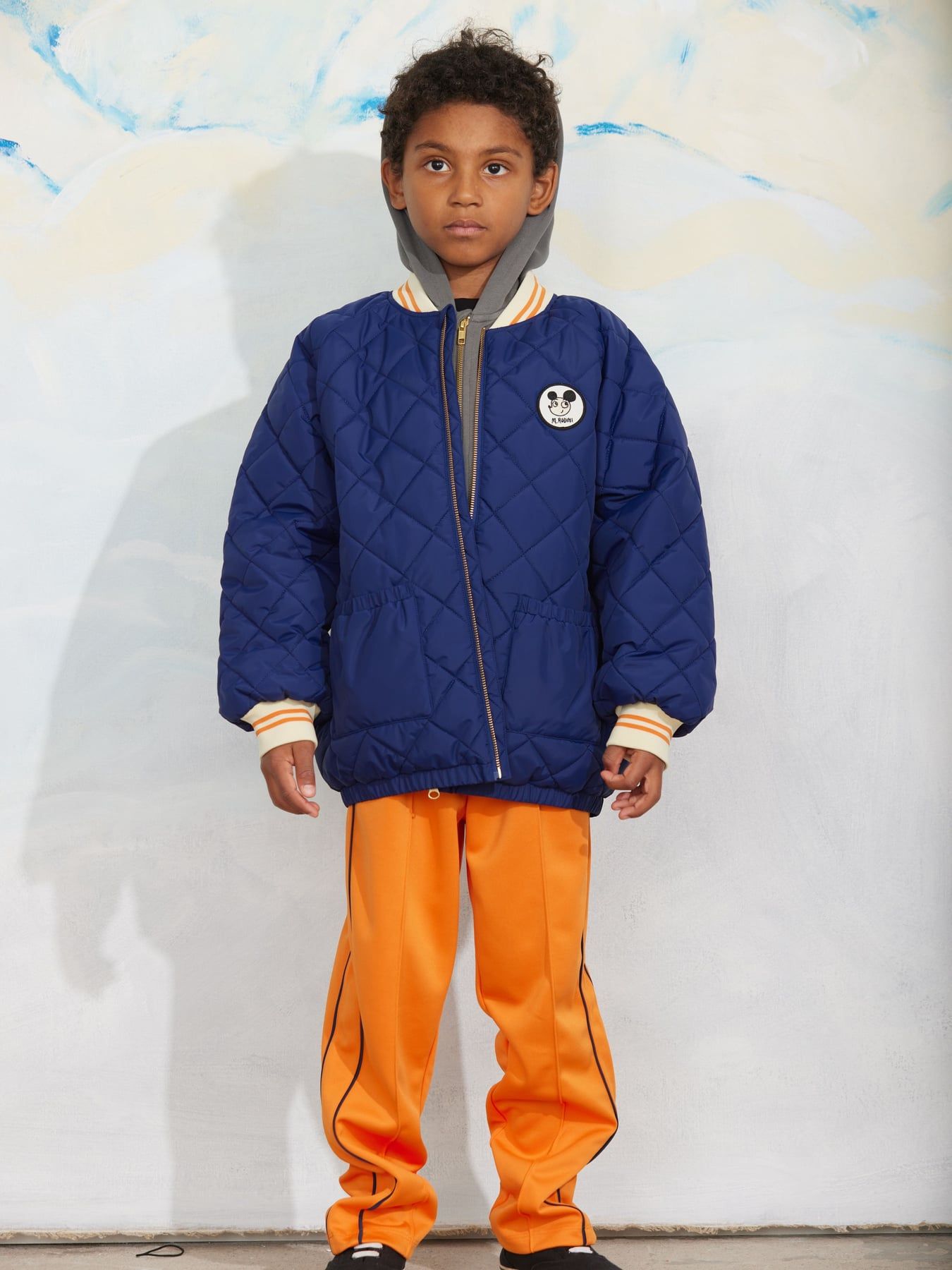 A child model wearing a navy blue quilted bomber jacket with a gray hood and orange track pants, standing against a textured white wall.