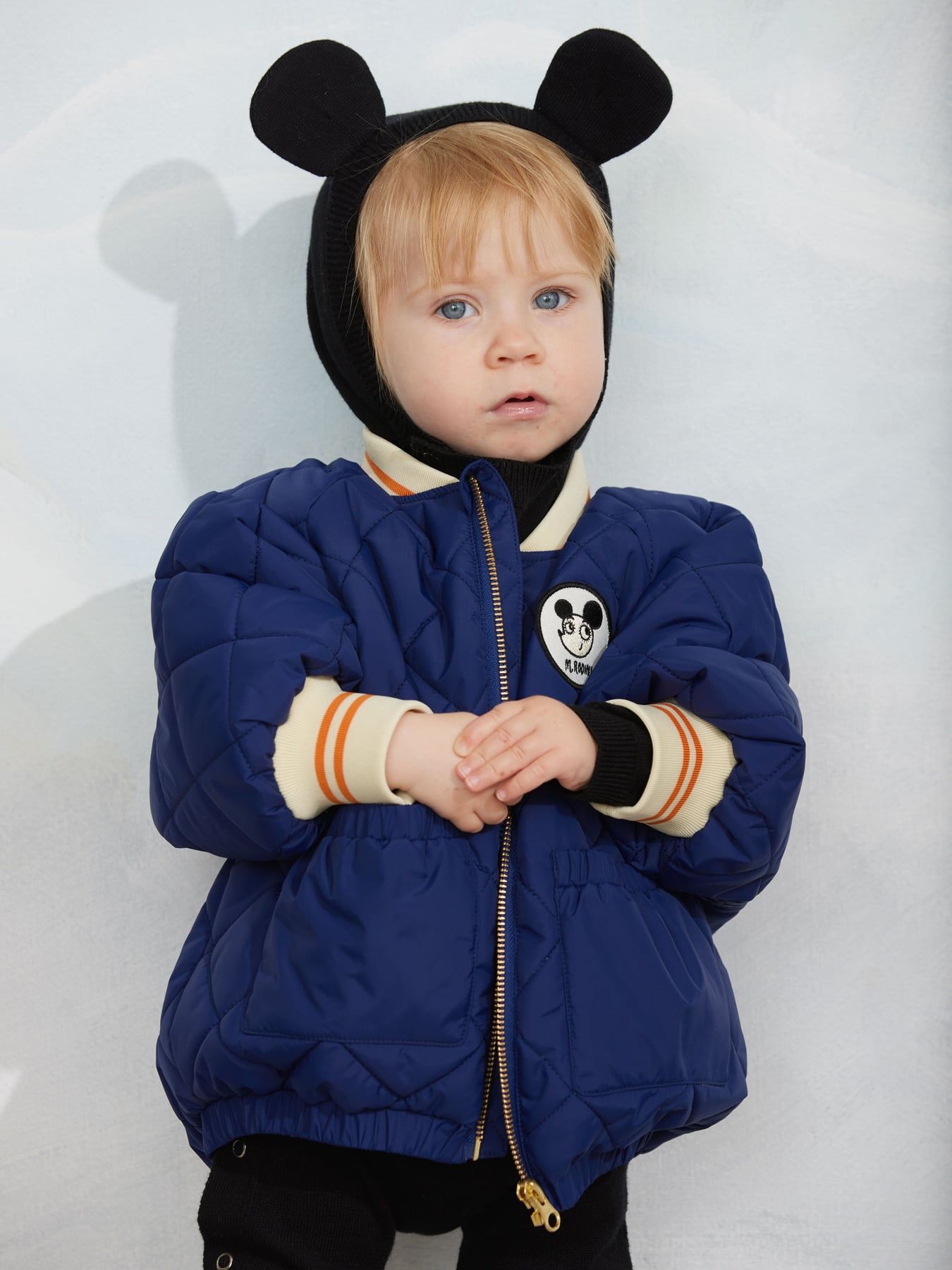 A toddler wearing a navy blue puffer jacket with a black hood featuring mouse ears, cream ribbed cuffs, and a Mickey Mouse patch on the chest, standing against a light gray background.