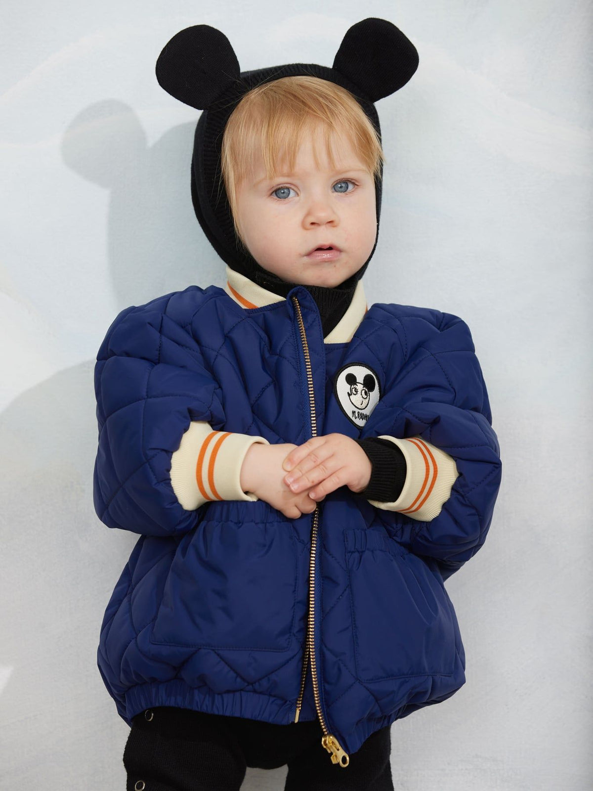 A toddler wearing a navy blue puffer jacket with a black hood featuring mouse ears, cream ribbed cuffs, and a Mickey Mouse patch on the chest, standing against a light gray background.